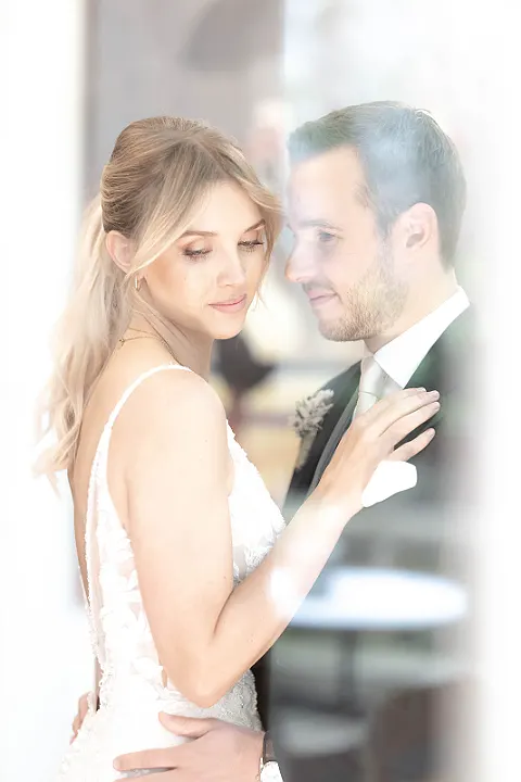 Ein Brautpaar posiert seitlich, die Braut in einem Hochzeitskleid mit tiefer Rückenpartie und der Bräutigam im Anzug.