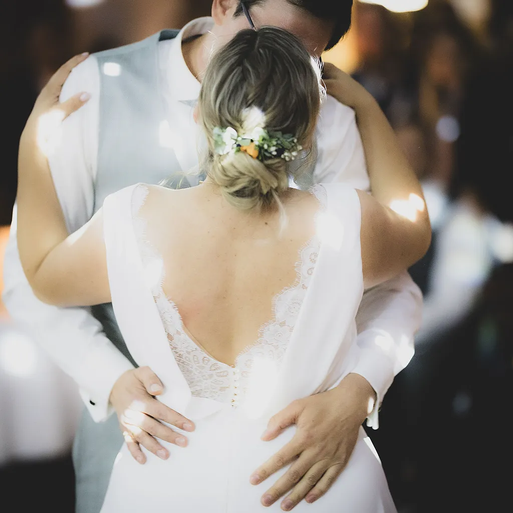 Ein Brautpaar umarmt sich, die Braut trägt ein weißes Kleid mit Spitzenrücken und Blumen im Haar.