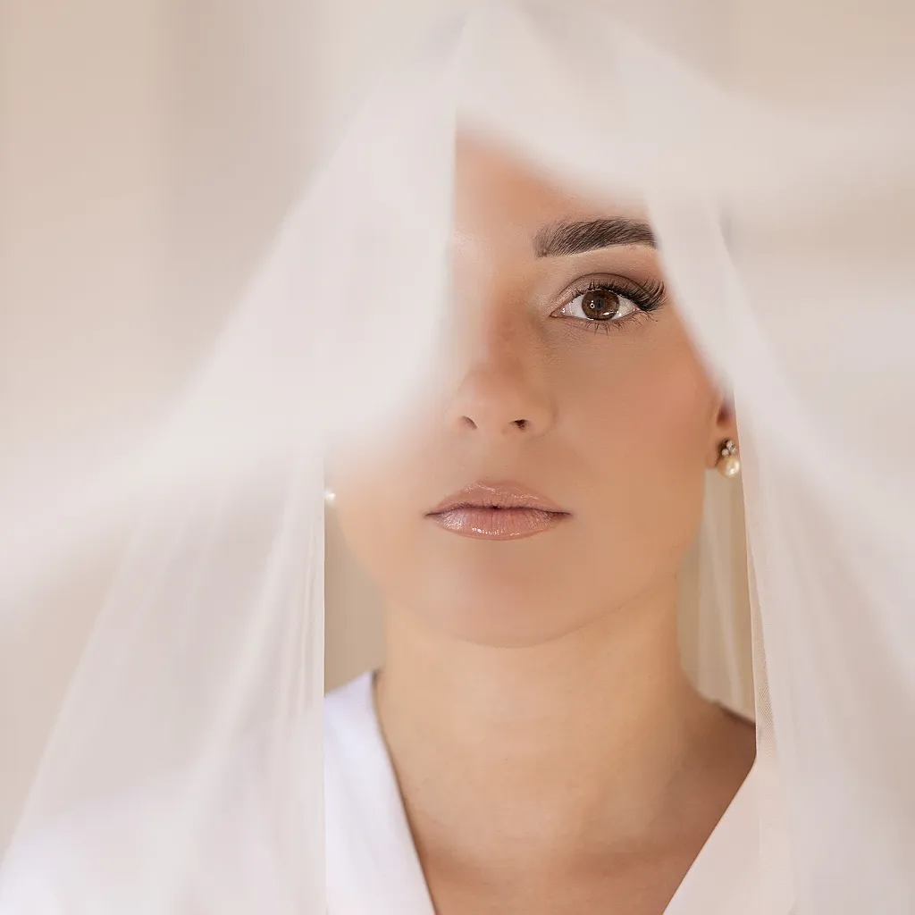 Nahaufnahme einer Person mit Brautschleier über dem Gesicht, ein Auge und Mund sind sichtbar.