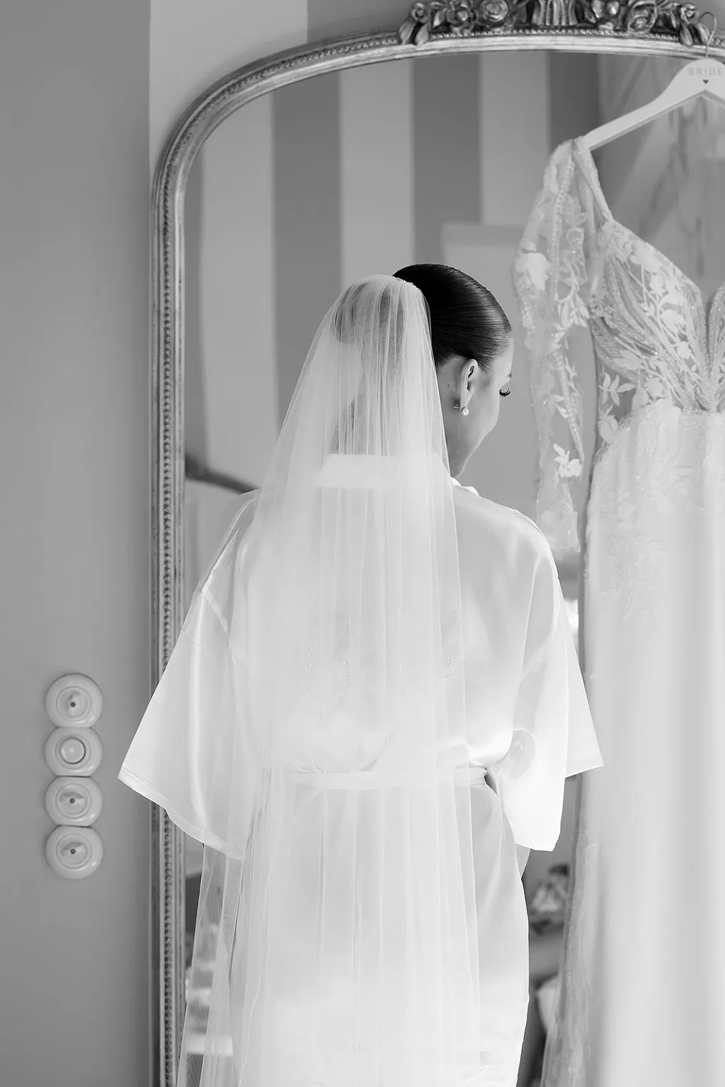Eine Person mit Schleier und Morgenmantel betrachtet ein Hochzeitskleid vor einem Spiegel. Schwarz-Weiß-Aufnahme.