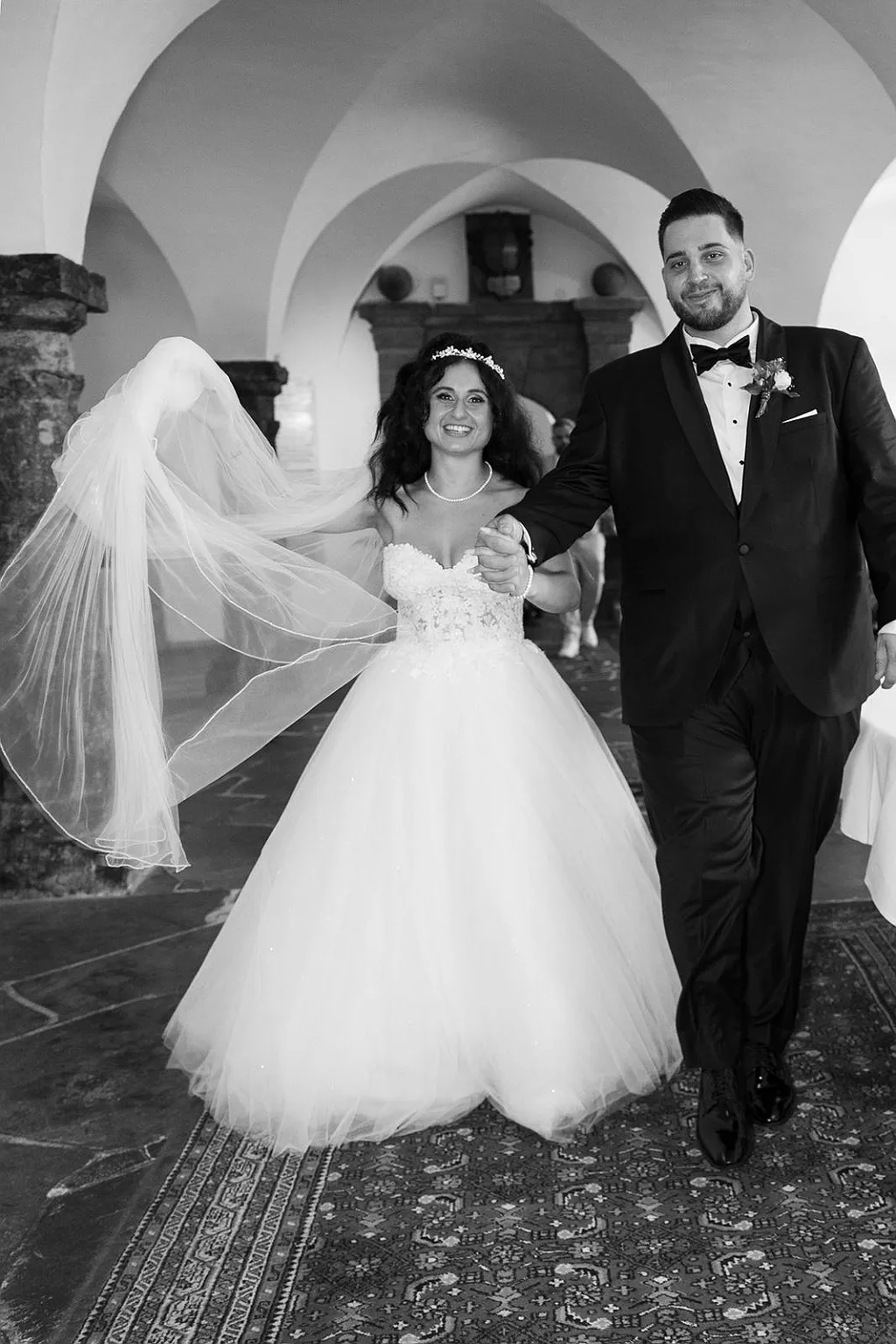 Brautpaar in Hochzeitskleidung läuft auf Teppich, im Hintergrund Gewölbe einer Halle.