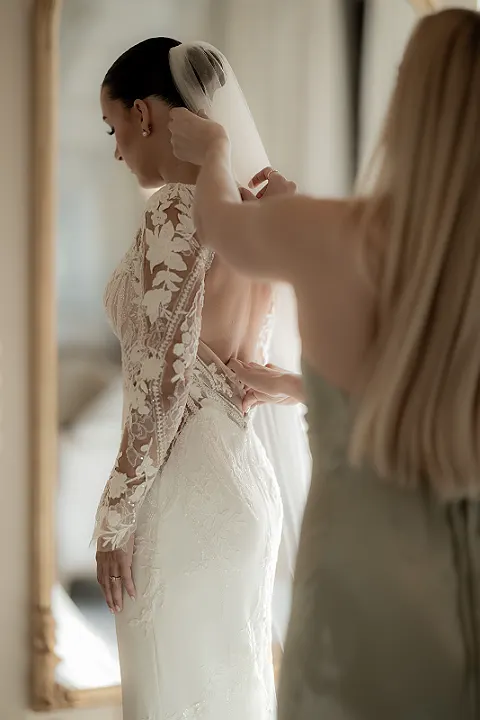 Eine Person in einem weißen Hochzeitskleid wird von einer anderen Person angekleidet. Das Kleid hat Spitzenärmel.