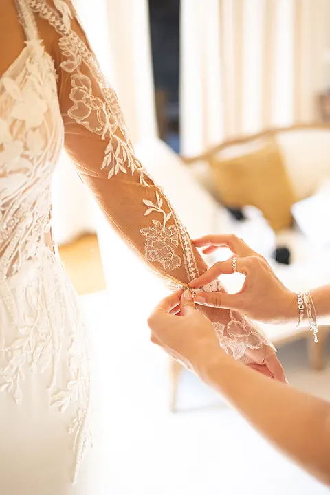Eine Person schließt Knöpfe an der Ärmelspitze eines bestickten Brautkleides. Das Kleid ist langärmlig.