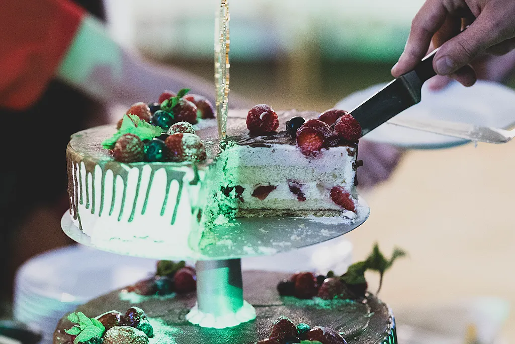 Eine Hand schneidet ein Stück einer zweistöckigen Hochzeitstorte mit Beeren, Schokolade und Minze an.