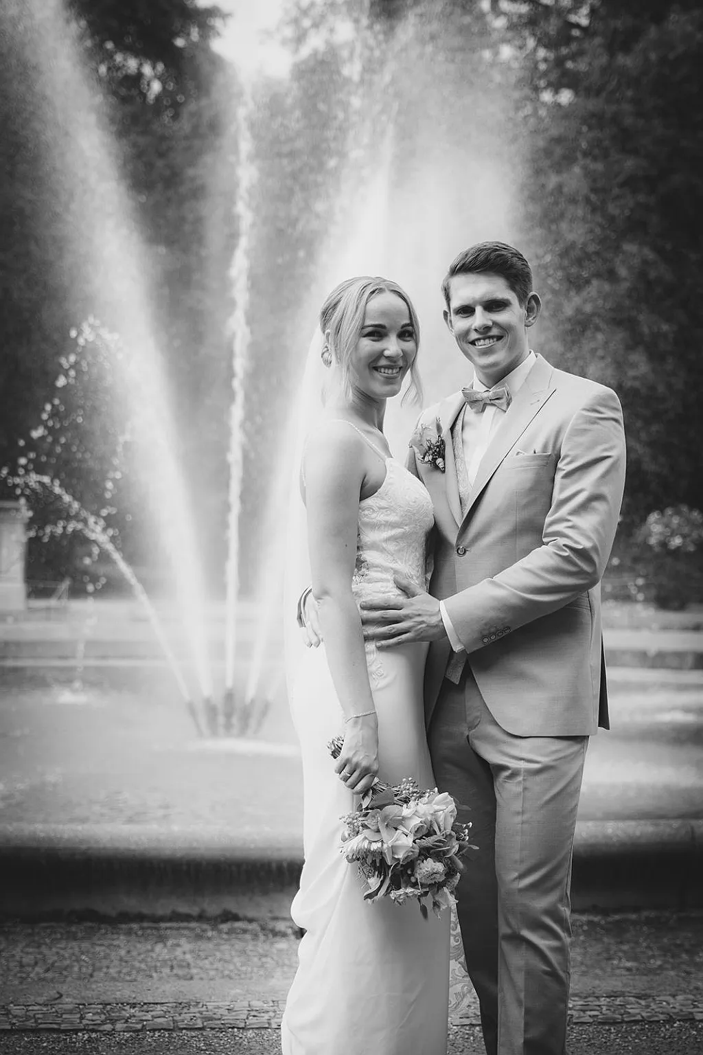Ein Brautpaar in Hochzeitskleidung lächelt vor einem Springbrunnen in Schwarz-Weiß.