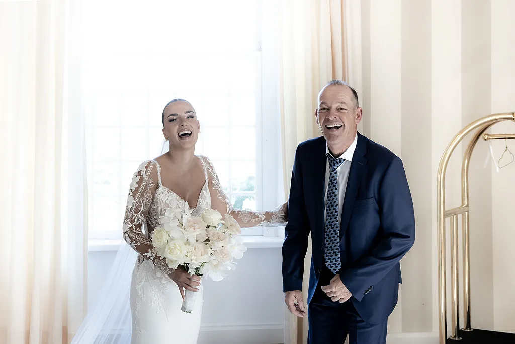 Braut im weißen Kleid mit Blumenstrauß, Bräutigam im dunklen Anzug, beide lachen vor einem Fenster.
