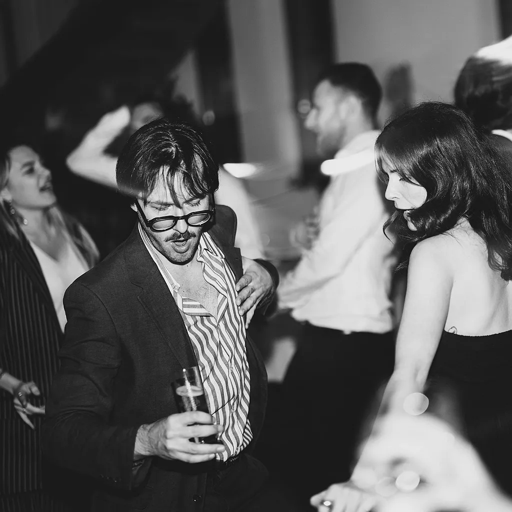 Ein Mann mit Brille und gestreiftem Hemd tanzt und hält ein Glas. Daneben tanzen weitere Gäste.