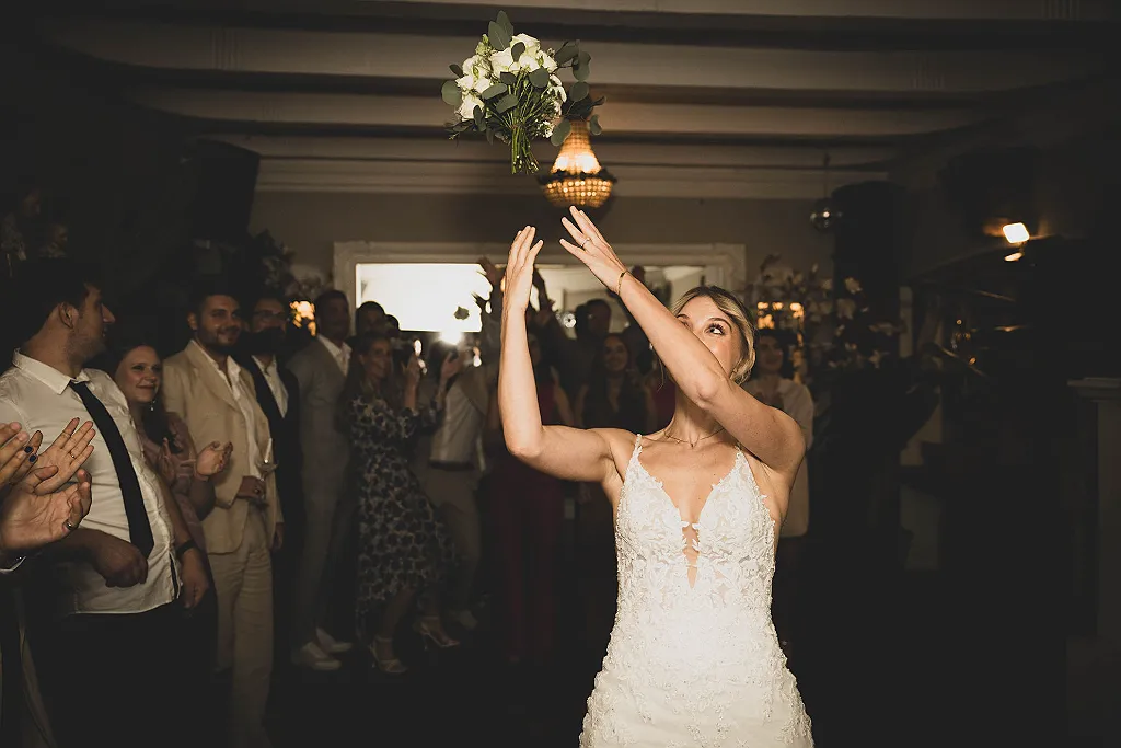 Eine Frau in einem weißen Hochzeitskleid wirft einen Brautstrauß in die Luft, im Hintergrund applaudieren Gäste.