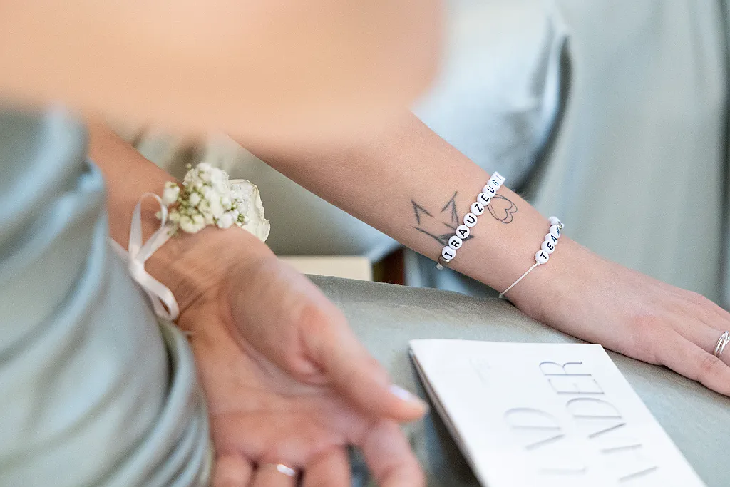 Arm einer Person mit Blumenarmband, Tattoo und Perlenarmband mit der Aufschrift "TRAUZEUGE TEAM".