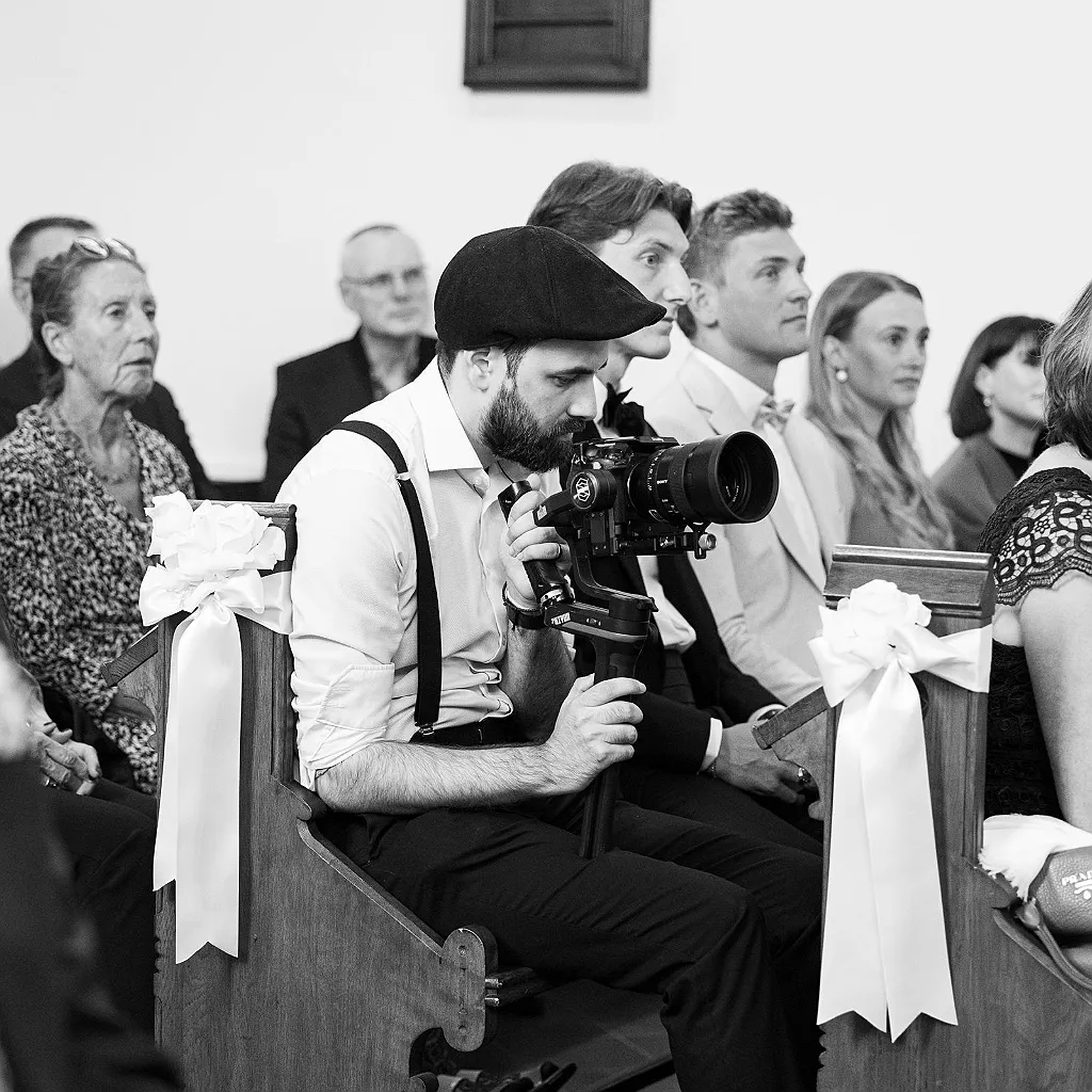 Ein Mann mit Bart und Schiebermütze filmt mit einer Kamera in einer Kirchenbank, umgeben von weiteren Gästen.