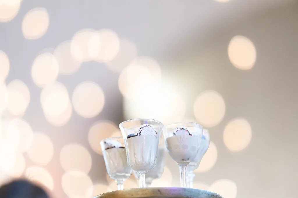 Mehrere Dessertgläser mit heller Creme und dunklem Topping stehen auf einem Tablett, im Hintergrund Bokeh-Lichter.