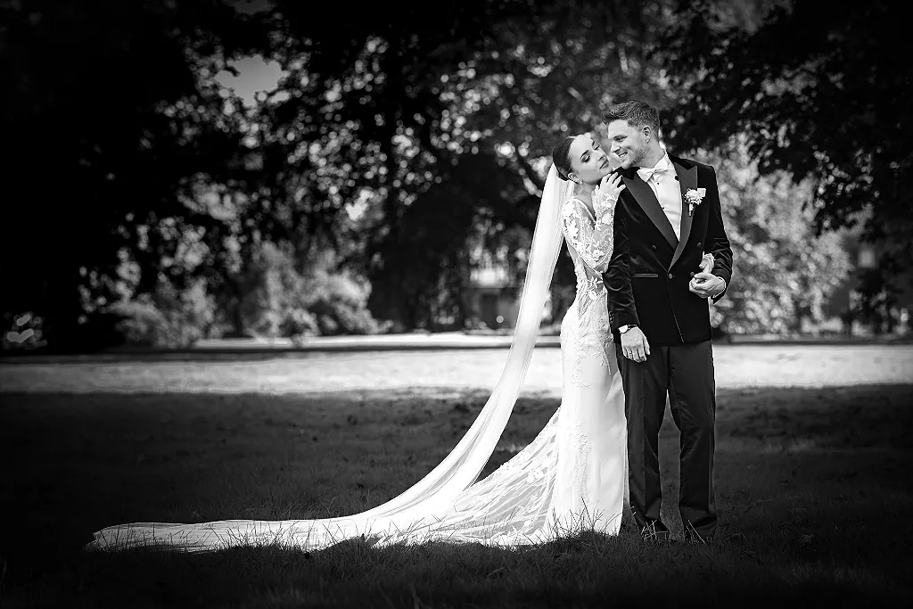 Brautpaar in Hochzeitskleidung steht auf einer Wiese vor Bäumen im Gegenlicht. Schwarz-Weiß-Aufnahme.