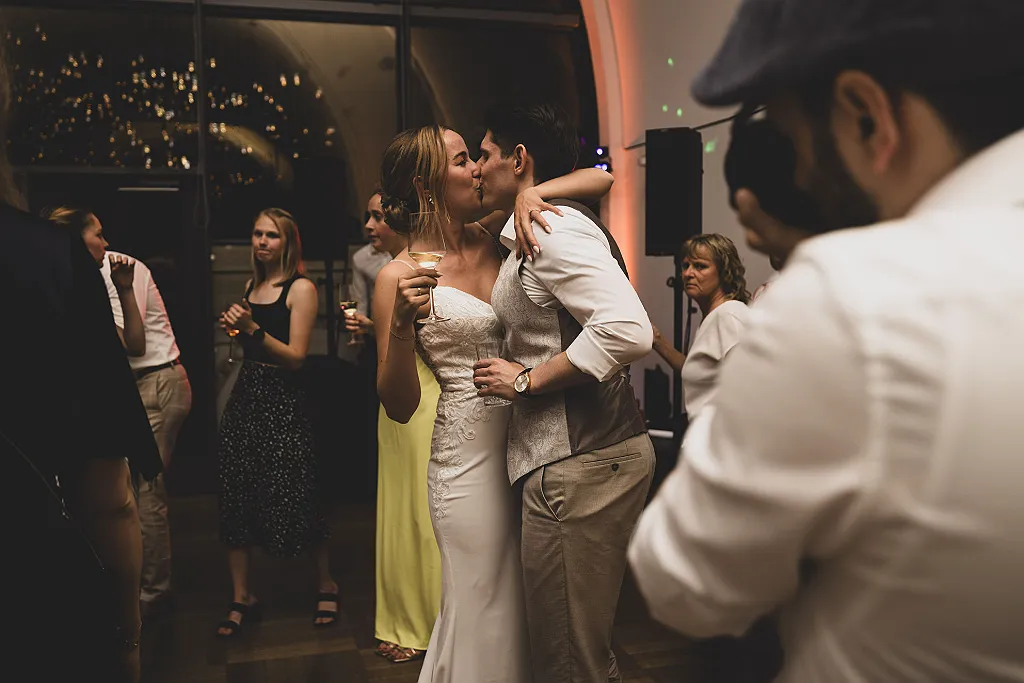 Ein Brautpaar küsst sich inmitten tanzender Gäste und feiert die Hochzeit bei gedämpftem Licht.