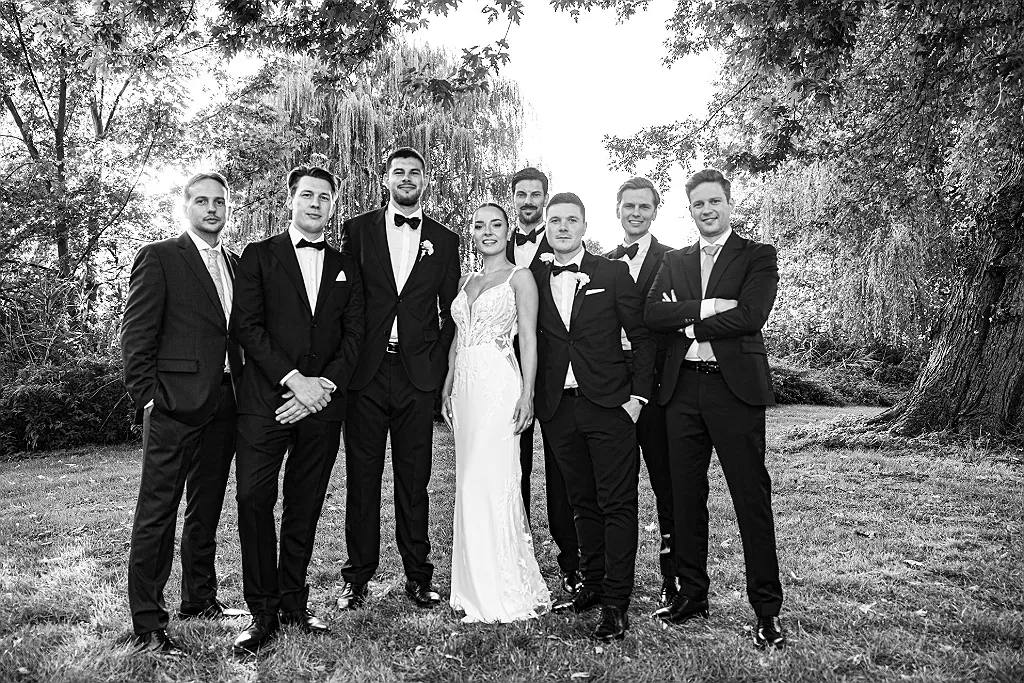 Eine Gruppe von sechs Männern in Anzügen und eine Frau im Brautkleid stehen auf einer Wiese vor Bäumen.
