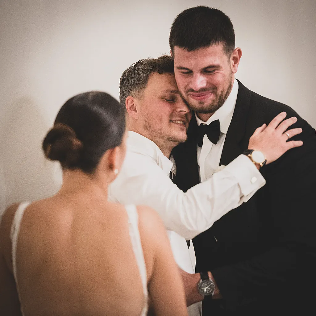 Zwei Männer umarmen sich, eine Frau mit Hochzeitskleid ist im Vordergrund unscharf zu sehen.