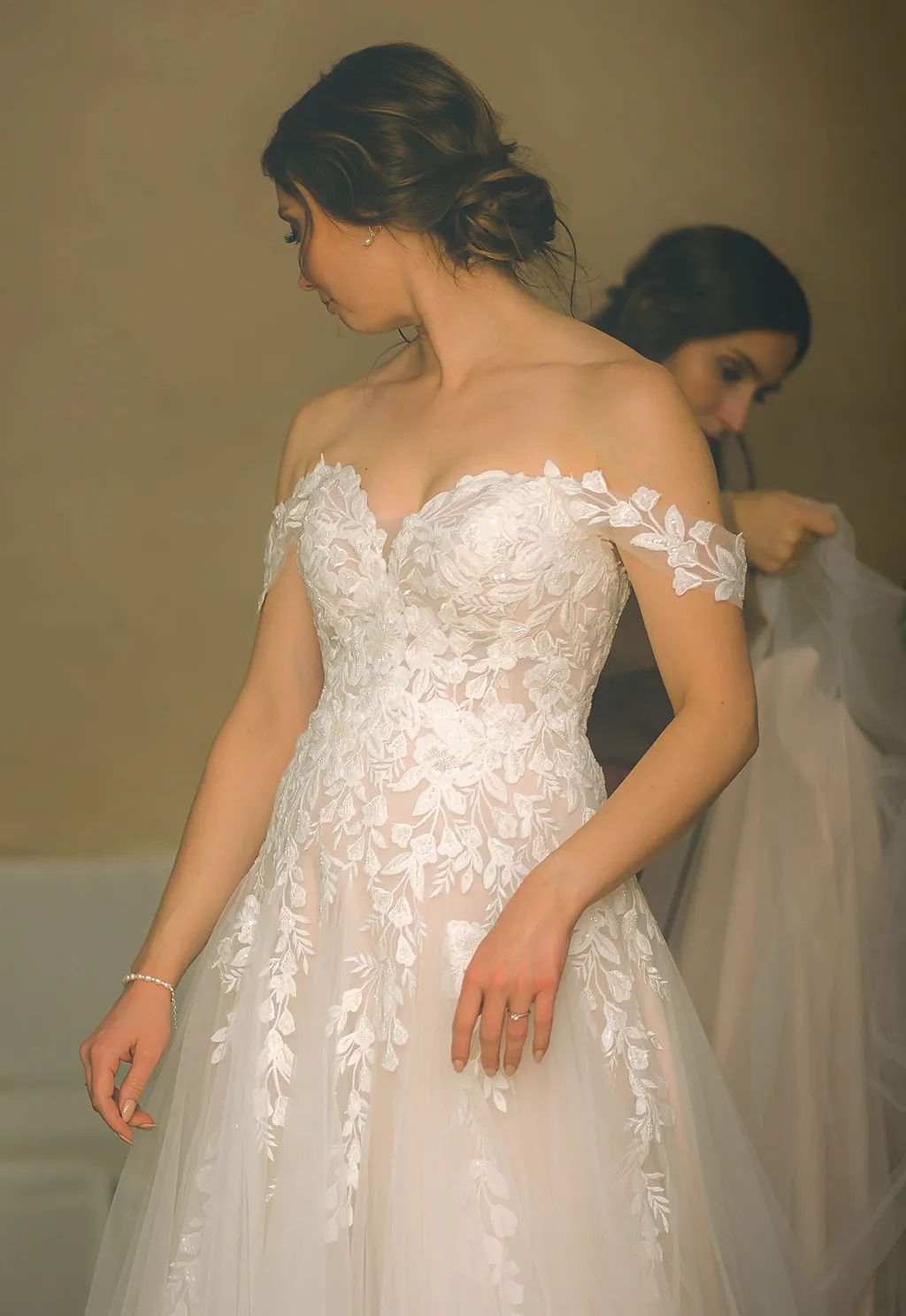 Eine Person in einem weißen Hochzeitskleid mit floralen Applikationen und einer weiteren Person im Hintergrund.