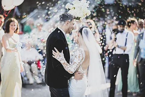 Brautpaar im Hochzeitsoutfit umarmt sich, umgeben von Seifenblasen, während Gäste im Hintergrund stehen.