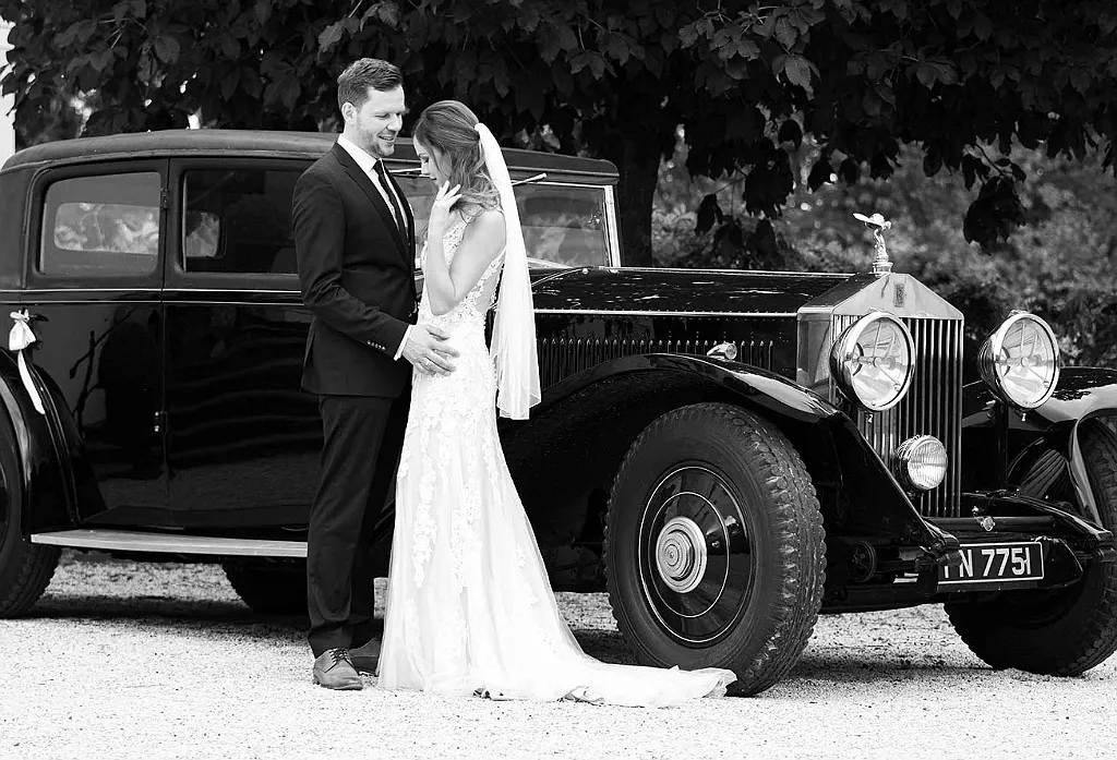 Brautpaar in Hochzeitskleidung, die neben einem schwarzen Oldtimer posieren, im Freien.