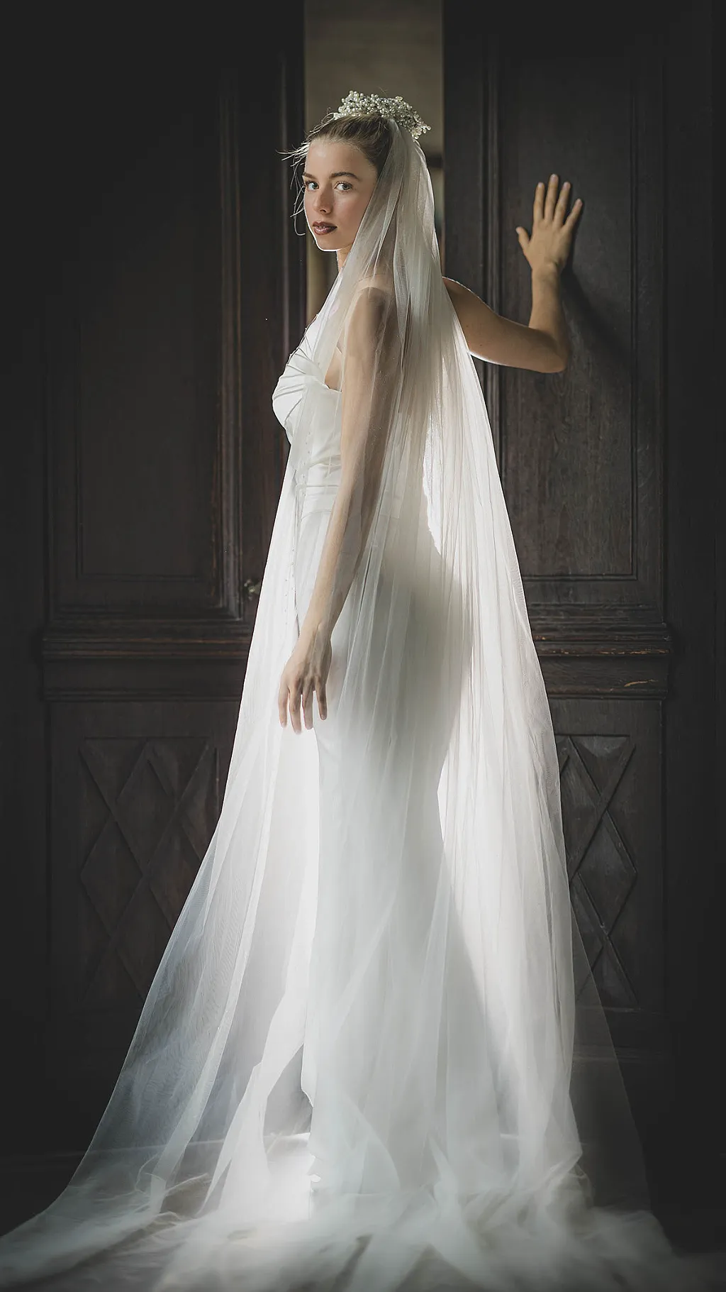 Eine Frau im weißen Brautkleid mit Schleier und Diadem steht vor einer dunklen Holztür.