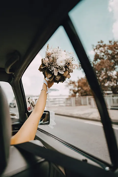 Ein Arm hält einen Hochzeitsstrauß aus dem Fenster eines fahrenden Autos. Der Strauß ist aus getrockneten Blumen.
