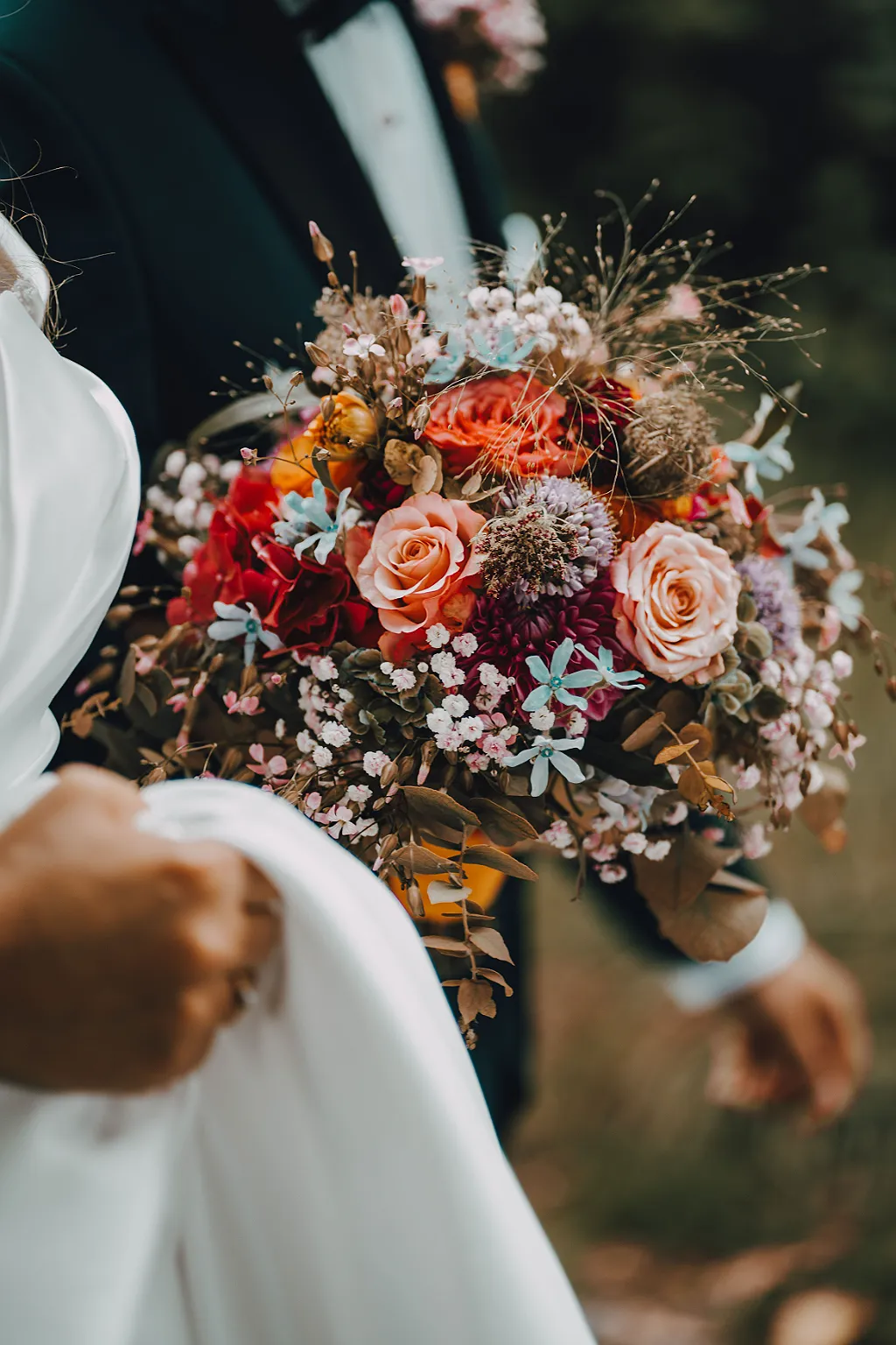 Großaufnahme eines Hochzeitsstraußes mit roten und rosa Rosen, blauen und weißen Akzenten, im Vordergrund ein weißes Kleid.