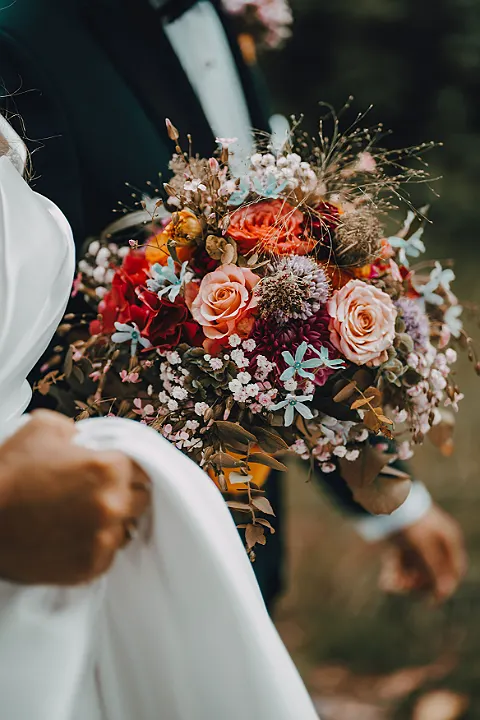 Großaufnahme eines Hochzeitsstraußes mit roten und rosa Rosen, blauen und weißen Akzenten, im Vordergrund ein weißes Kleid.