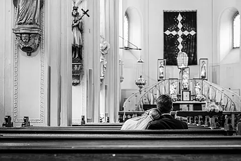 Rückenansicht eines Paares in einer Kirchenbank, Blick auf den Altarraum mit Kreuz und Statuen in Schwarz-Weiß.