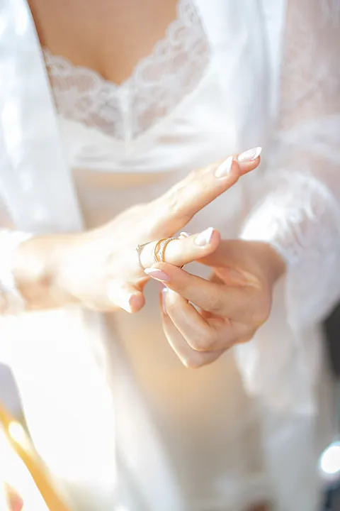 Eine Person schiebt einen goldenen Ring auf einen Finger, trägt ein weißes Spitzenoberteil und einen Morgenmantel.