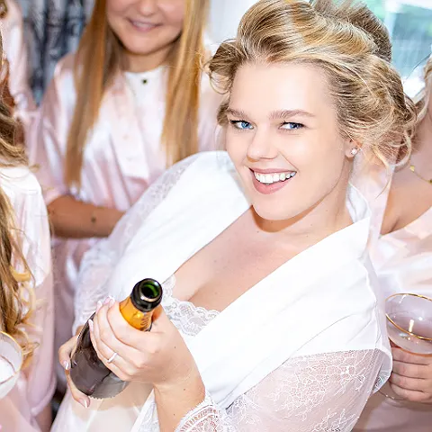 Eine Frau mit Lockenwicklern auf dem Kopf und Robe öffnet eine Flasche Sekt.