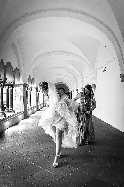 Eine Person in einem Hochzeitskleid beugt sich in einem Gang nach vorne, eine zweite Person stützt sie.