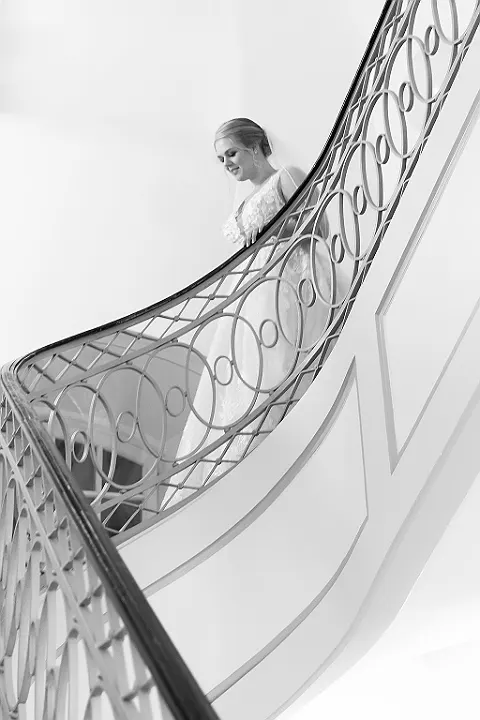 Eine Braut steht auf einer geschwungenen Treppe mit Geländer, aufgenommen aus der oberen Perspektive in Schwarz-Weiß.