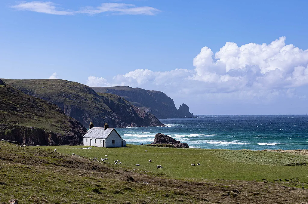 Ein weißes Haus am Meer mit grünen Hügeln und Schafen unter blauem Himmel mit Wolken.