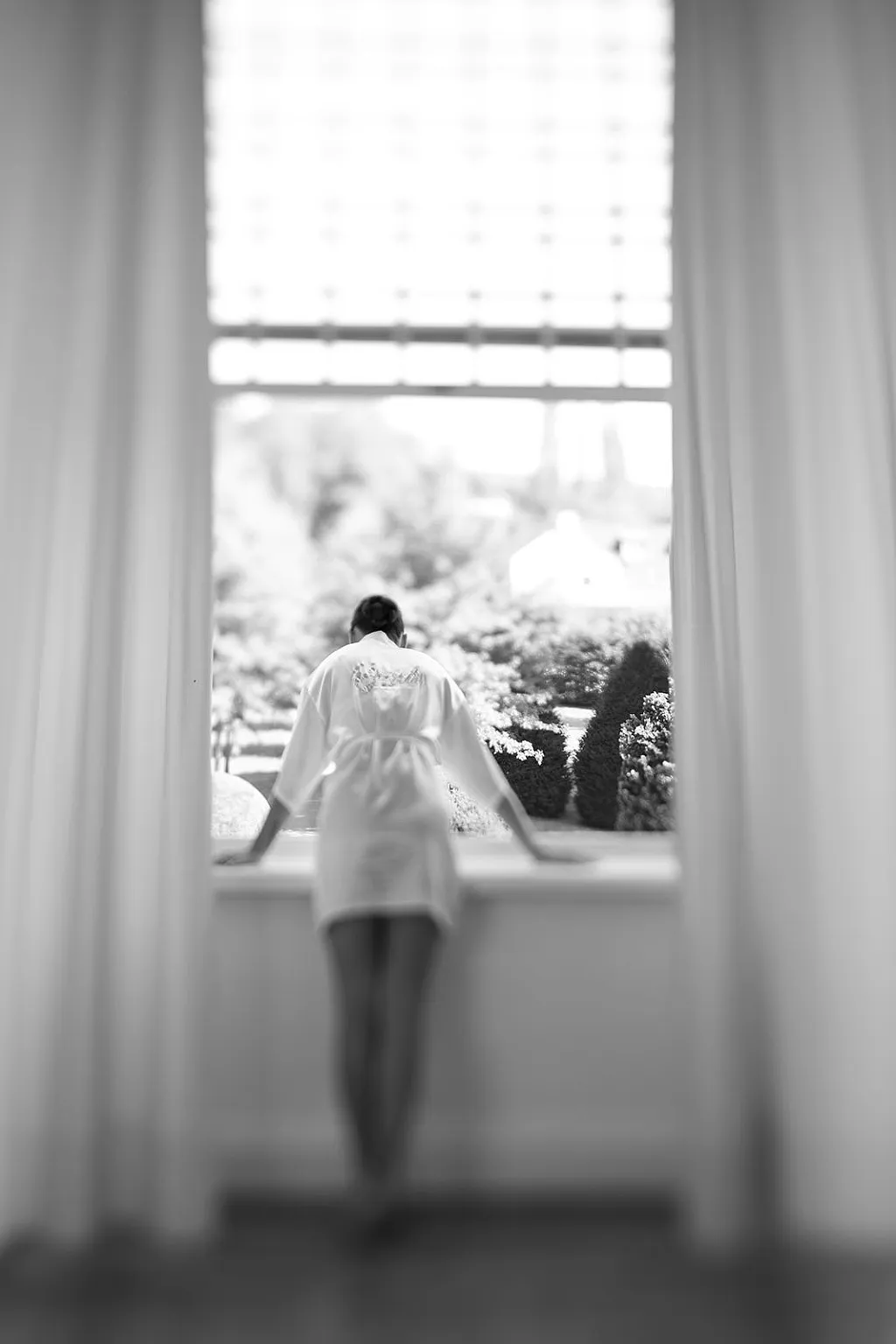 Eine Person in einem Bademantel blickt aus einem Fenster in einen grünen Garten. Die Vorhänge sind offen.