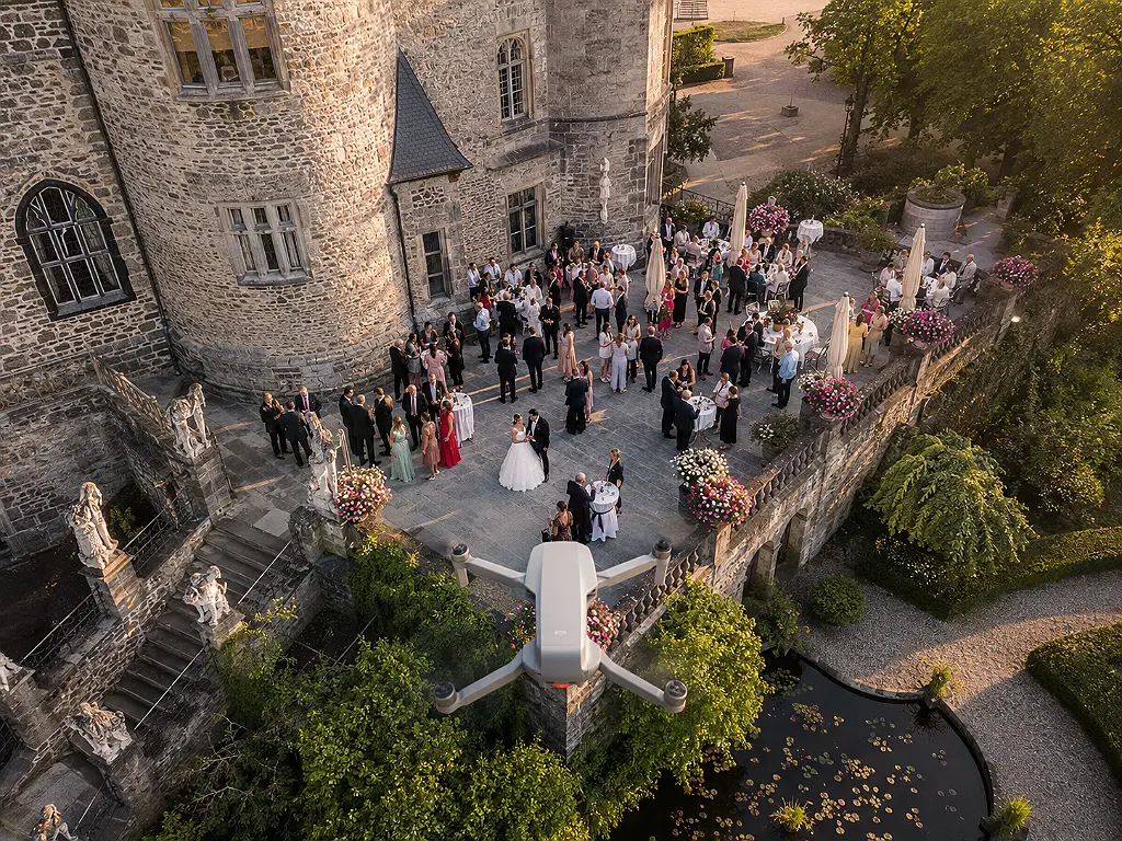 Drohnenaufnahme einer Hochzeitsgesellschaft auf einer Terrasse vor einem Schloss, inklusive einer Drohne im Vordergrund.